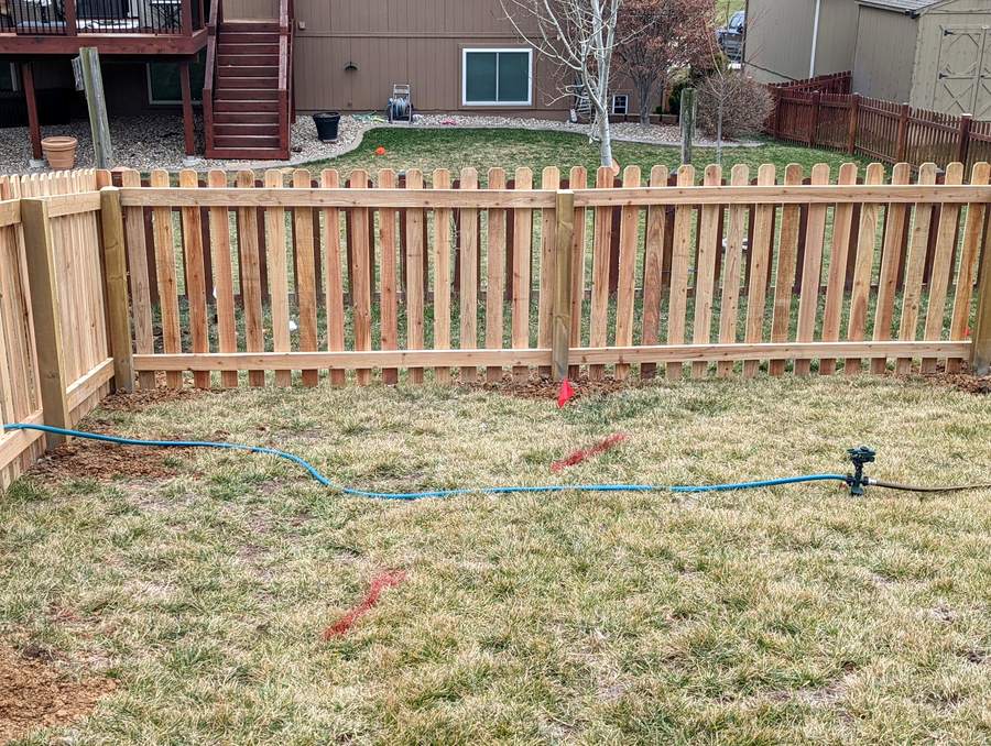 A spaced cedar picket fence with dog-ear tops running across a back yard below a raised deck.