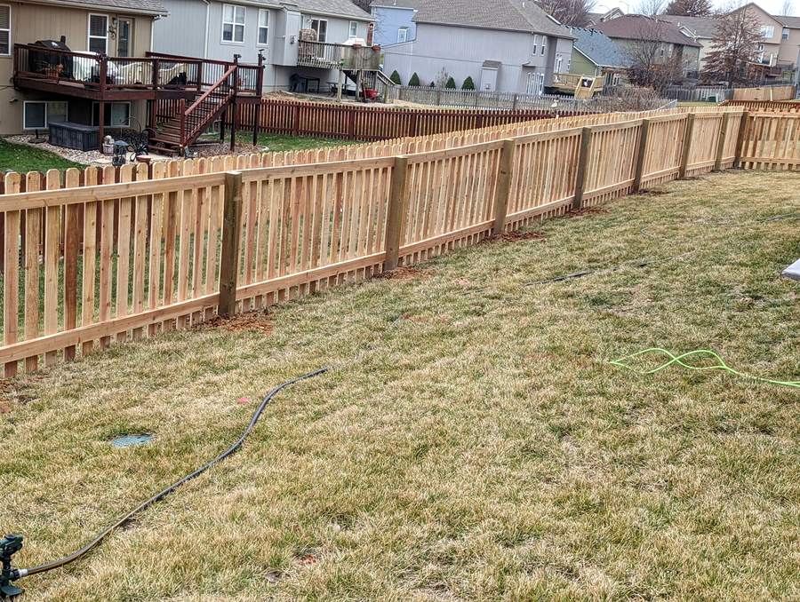 A long cedar picket fence running across the back of a mowed property with houses behind it.