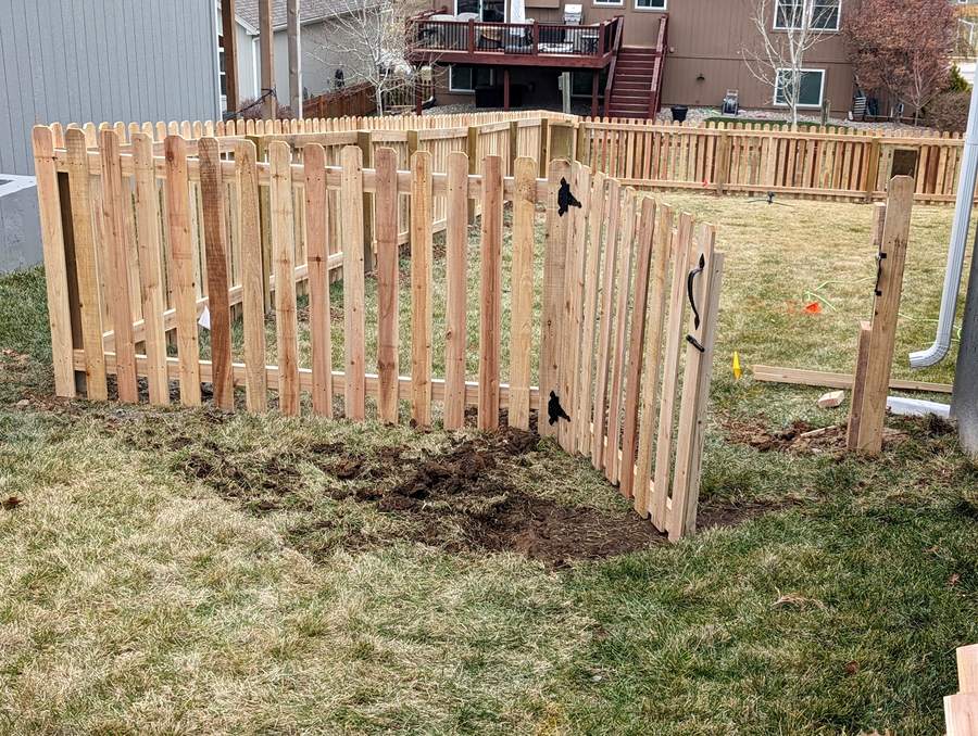A shadowbox picket gate standing open where a new fence line turns an angled corner, an ornate black pull fitted on the gate face.