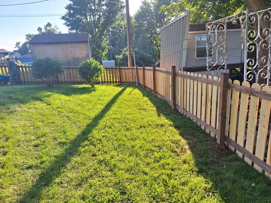 A cedar picket fence running down the edge of a mowed lawn beside a house.
