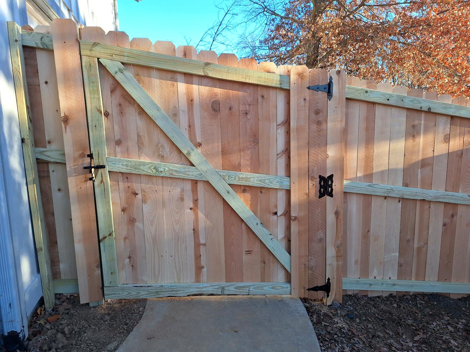 A finished cedar double gate with one leaf swung open, showing the vertical planks on the front and the two by four frame and diagonal brace on the back, hung on an antique strap hinge with an ornate pull plate and a drop rod latch.